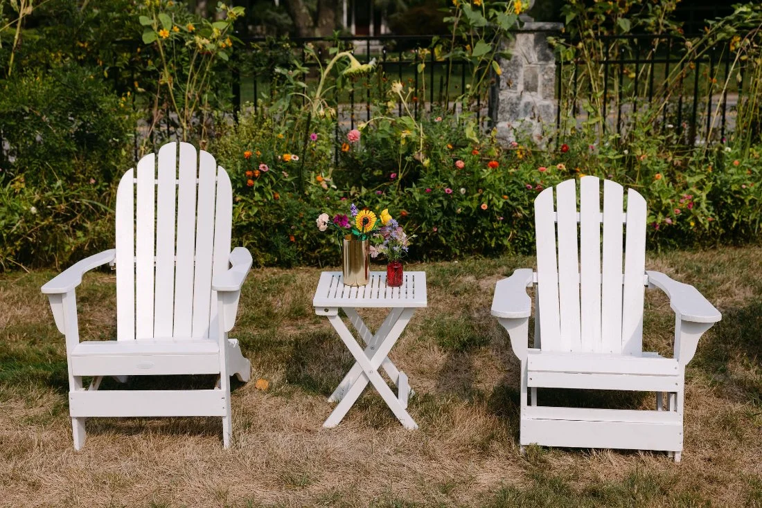 White Adirondack chairs and small table with flowers in garden lounge area at outdoor wedding in New York