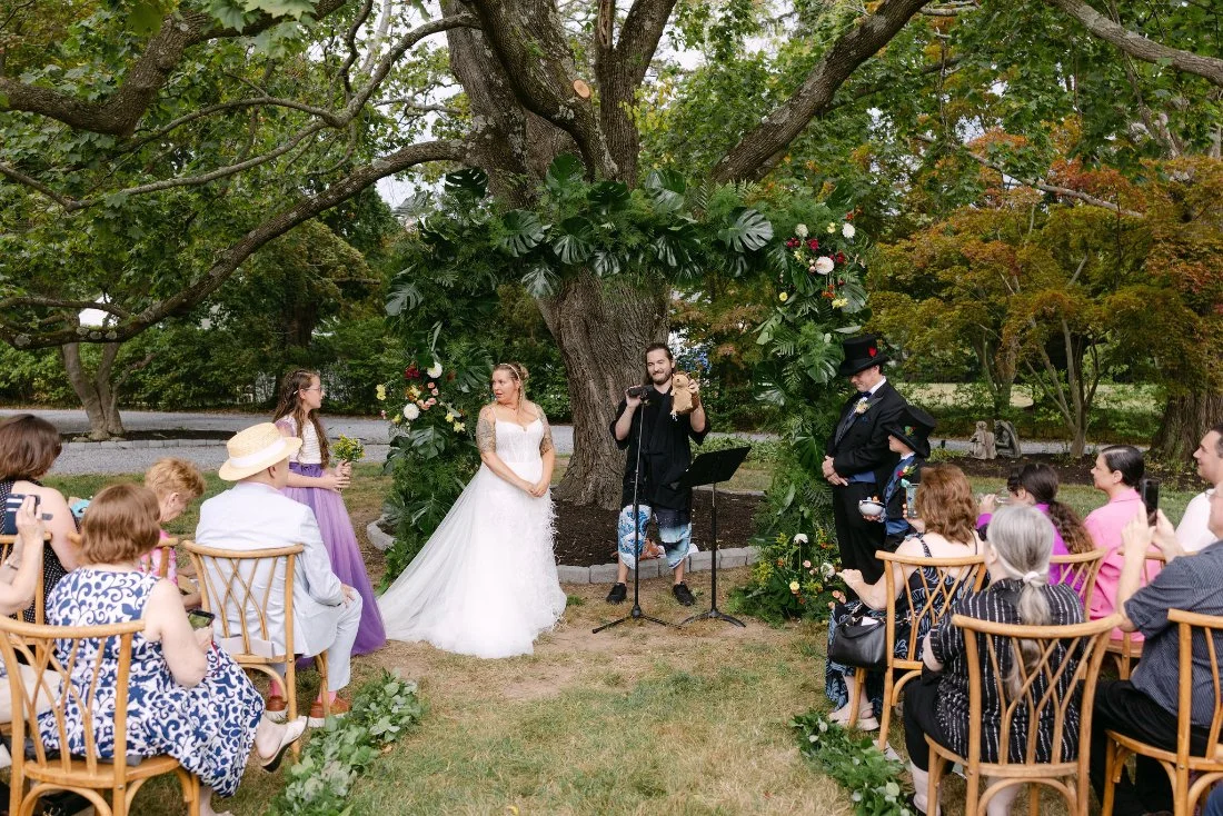 Bride, groom, officiant, and guests gathered under a large tree for heartfelt outdoor wedding ceremony in New York
