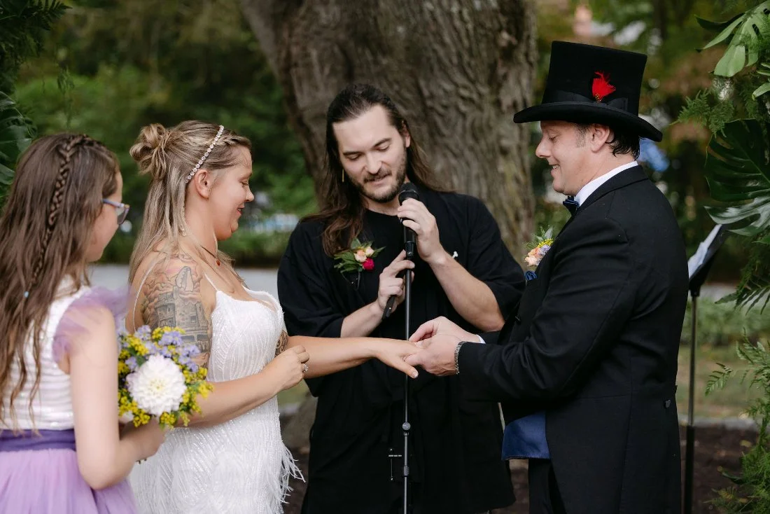 Bride and groom exchanging rings under the trees with their daughter by their side during outdoor wedding in New York