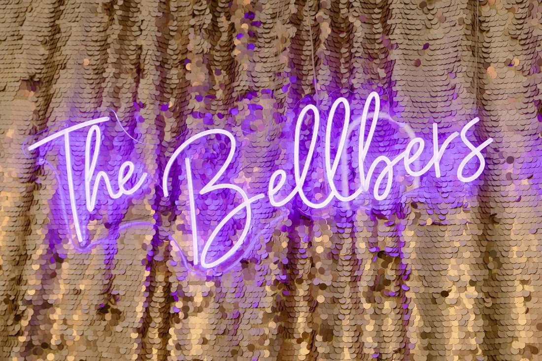 Custom “The Bellbers” neon sign on gold sequin backdrop at outdoor wedding in New York