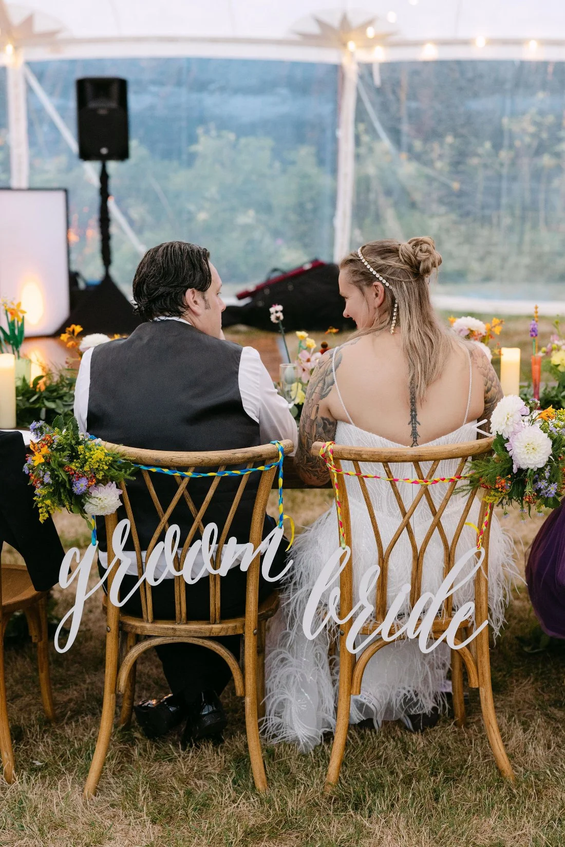 Bride and groom seen from behind, sitting at decorated chairs labeled 'bride' and 'groom' at their outdoor wedding in New York