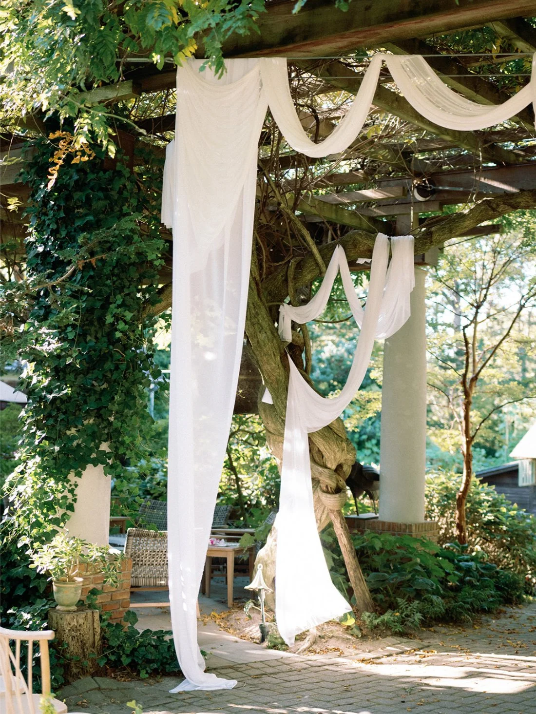 Flowing white fabric draped over a garden pergola during a Hamptons wedding ceremony setup.