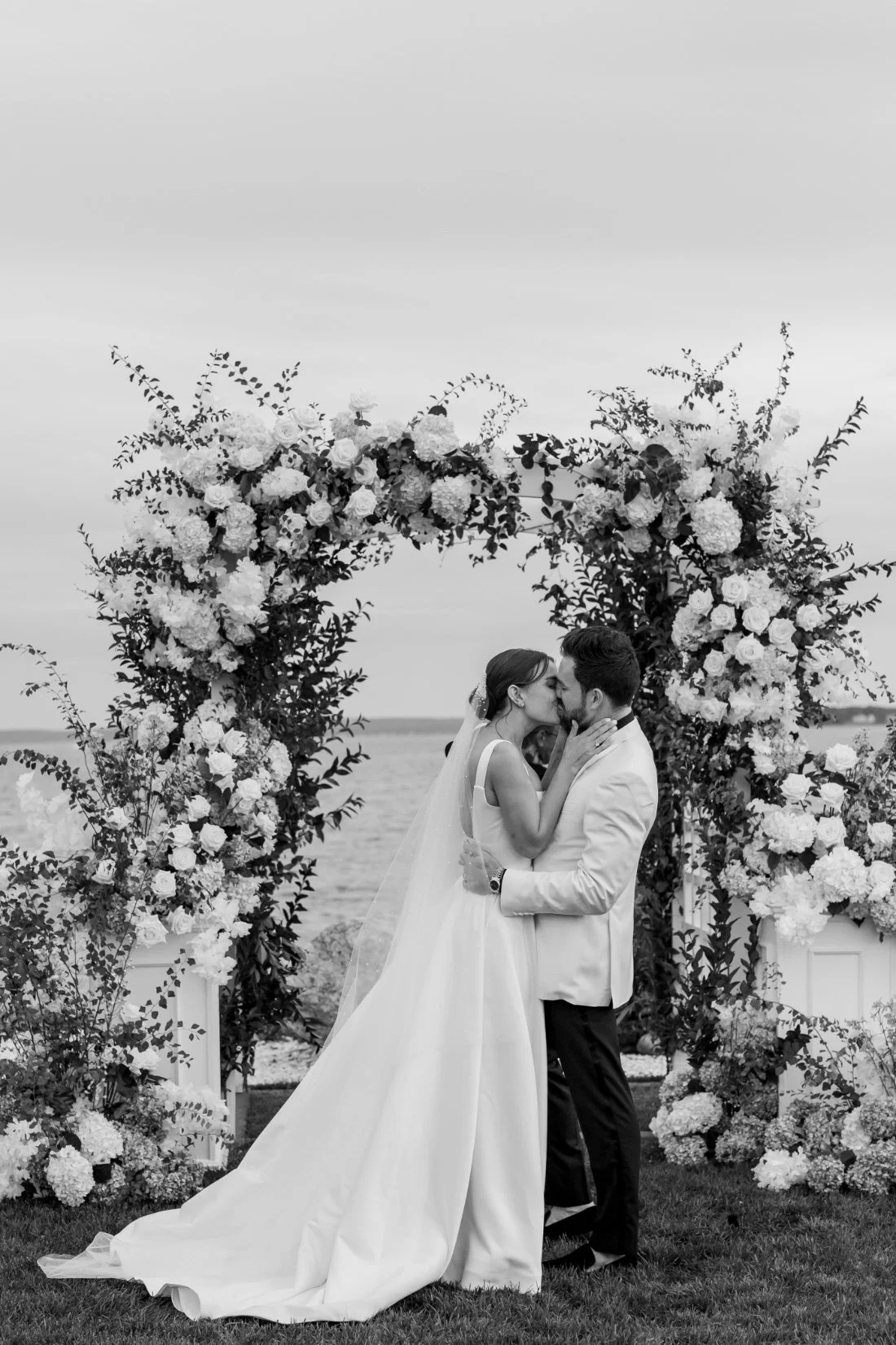 Black and white portrait of bride and groom kissing under floral arch at Peconic Bay Yacht Club