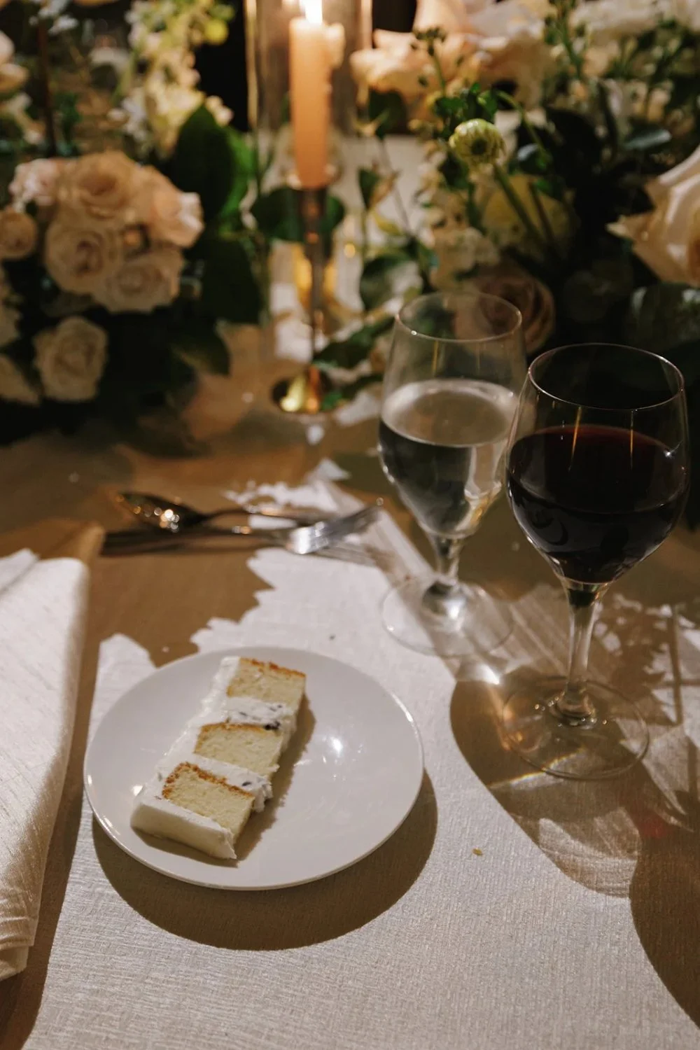 Wedding cake slices served on plate beside wine and candlelit centerpiece
