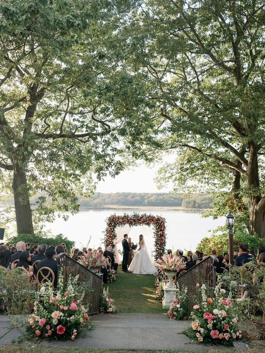 Guests watch the couple exchange vows beneath a floral chuppah overlooking the Long Island Sound during a romantic Wainwright House wedding in Rye, New York.