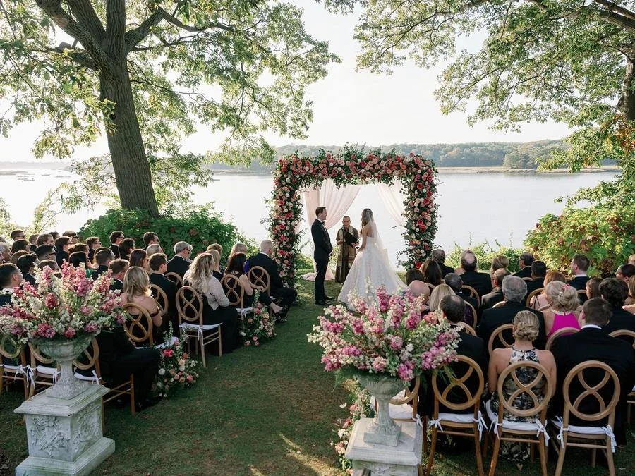 Guests gather beneath towering trees for a waterfront Wainwright House wedding ceremony with sweeping water views.