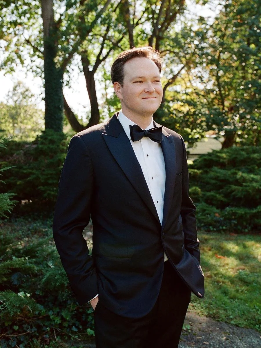 Groom portrait in a classic black tuxedo surrounded by lush greenery on the wedding day.