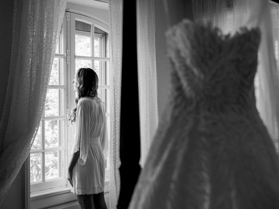 Bride stands by a sunlit window in her bridal robe while her wedding gown hangs nearby.