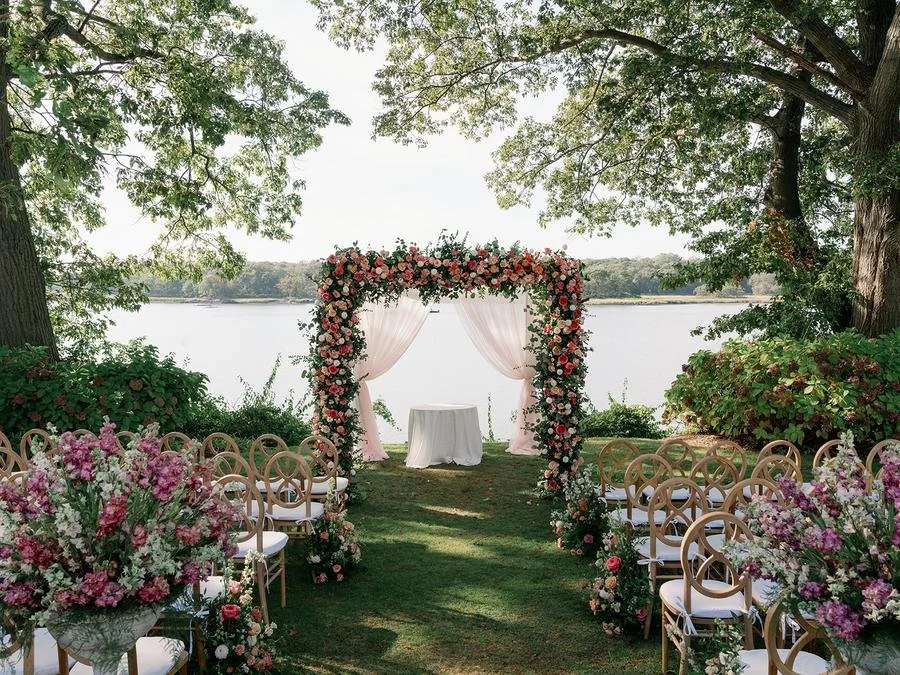 Waterfront ceremony setup with a floral arch and garden chairs for a Wainwright House wedding ceremony.