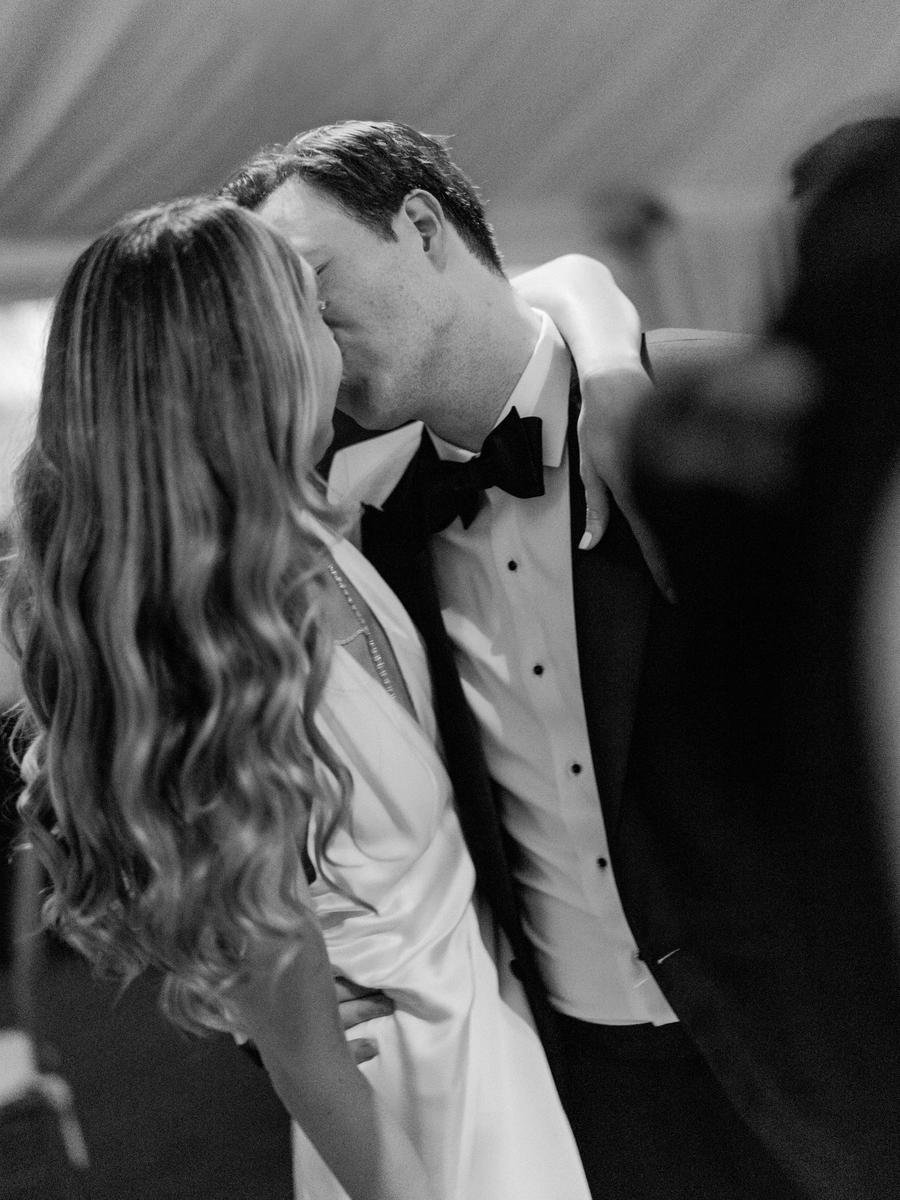 Black-and-white candid photo of the newlyweds sharing a kiss during the wedding reception.