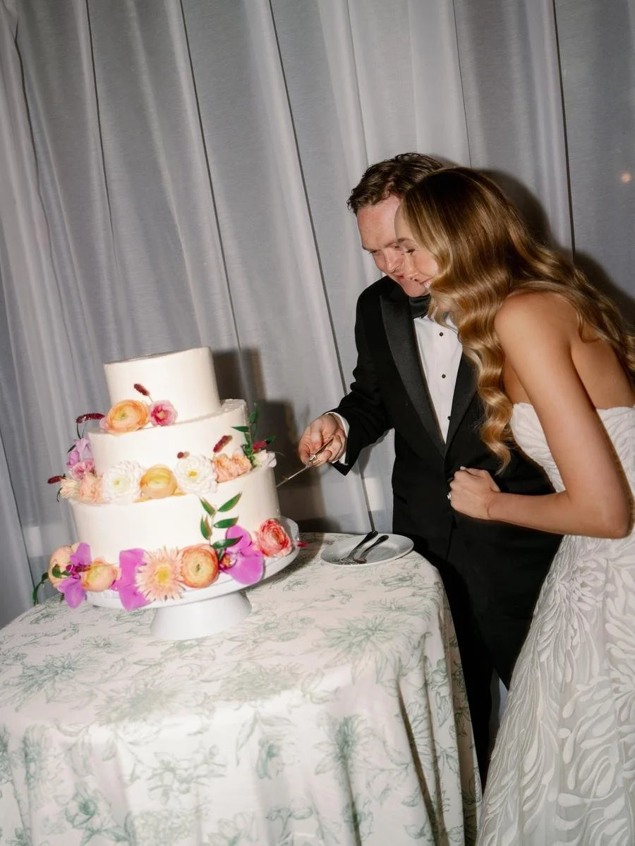 Bride and groom cutting their floral-adorned wedding cake during a joyful reception moment beneath draped tenting.