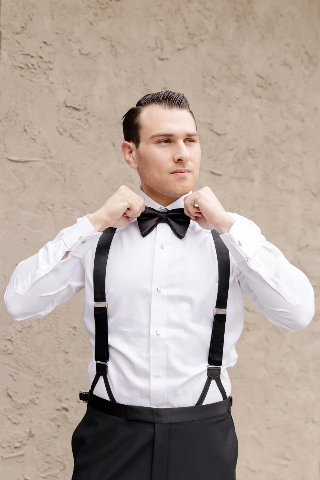 Groom adjusting his black bow tie and suspenders while getting ready for a classic New York wedding at Quaker Ridge Golf Club.