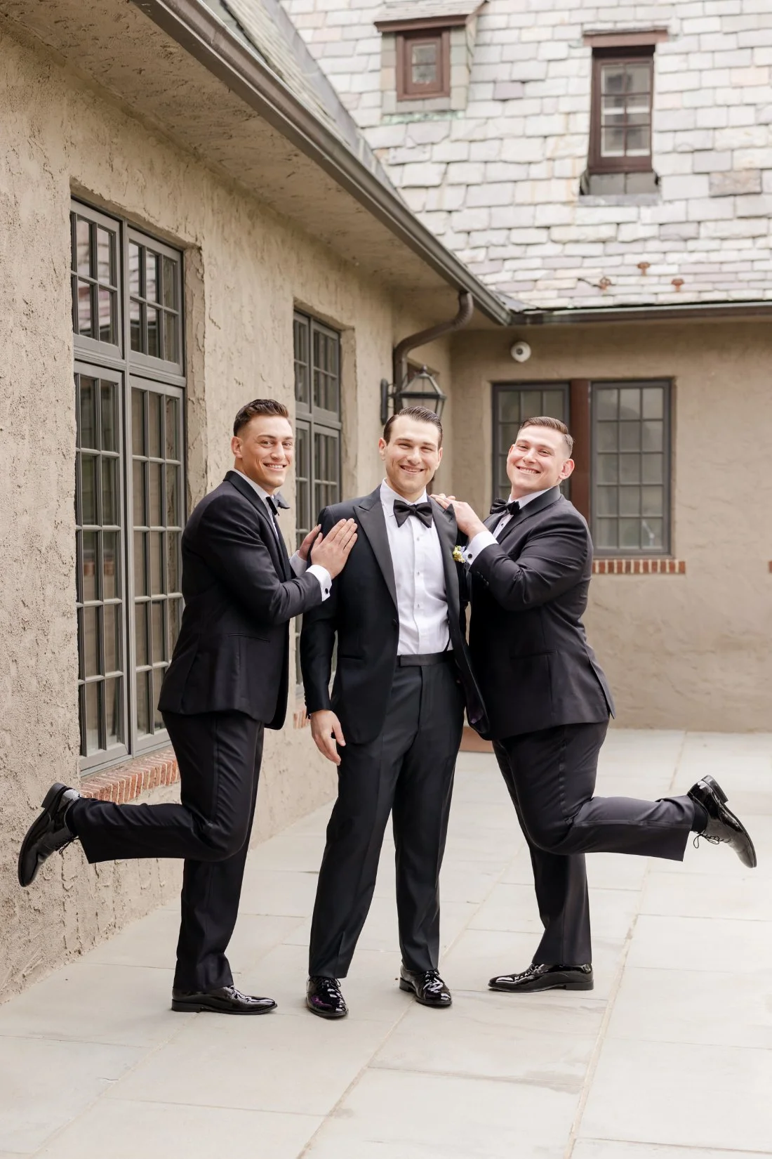 Groom posing outside with two groomsmen in classic black tuxedos before a Quaker Ridge Golf Club wedding.