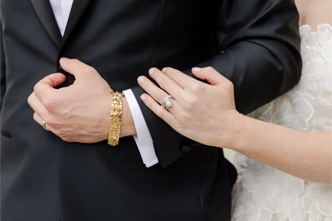 Close-up of bride’s wedding rings beside groom’s gold bracelet and black tuxedo