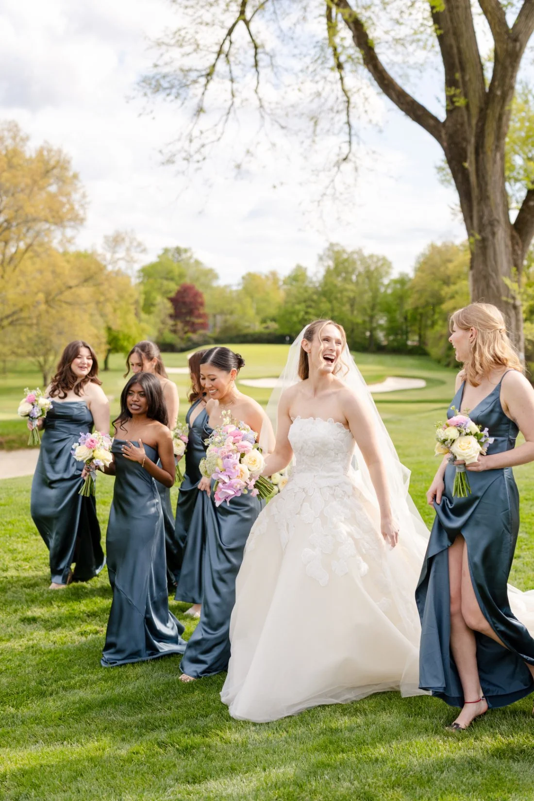 Bride laughs with bridesmaids carrying colorful spring bouquets on the golf course