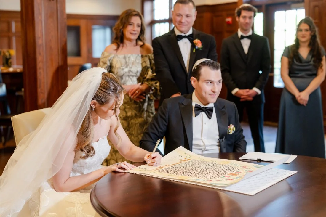 Bride signs the Ketubah as the groom and family look on during their Quaker Ridge Golf Club wedding.