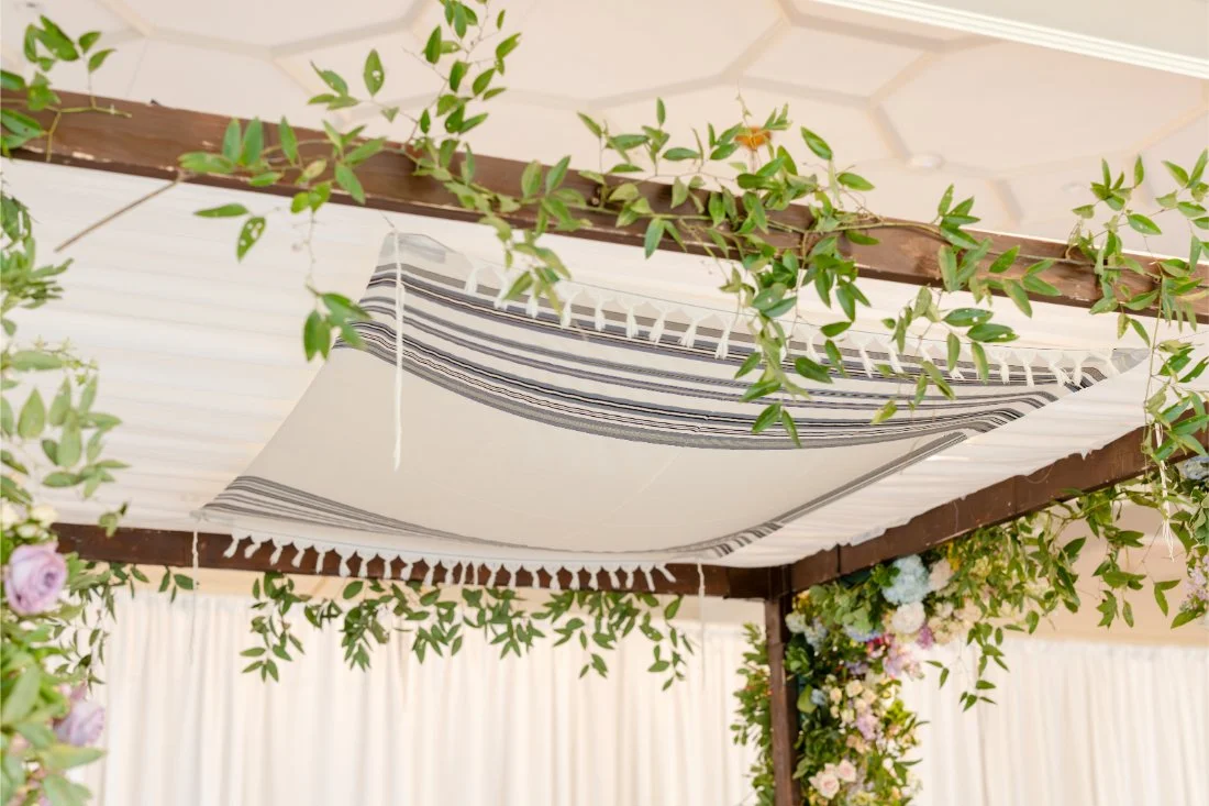 Traditional tallit draped beneath a greenery-covered chuppah for the Jewish wedding ceremony.