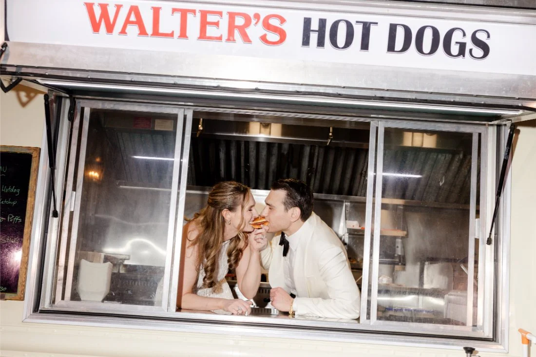 Bride and groom sharing a playful bite of a hot dog from the late-night food truck.
