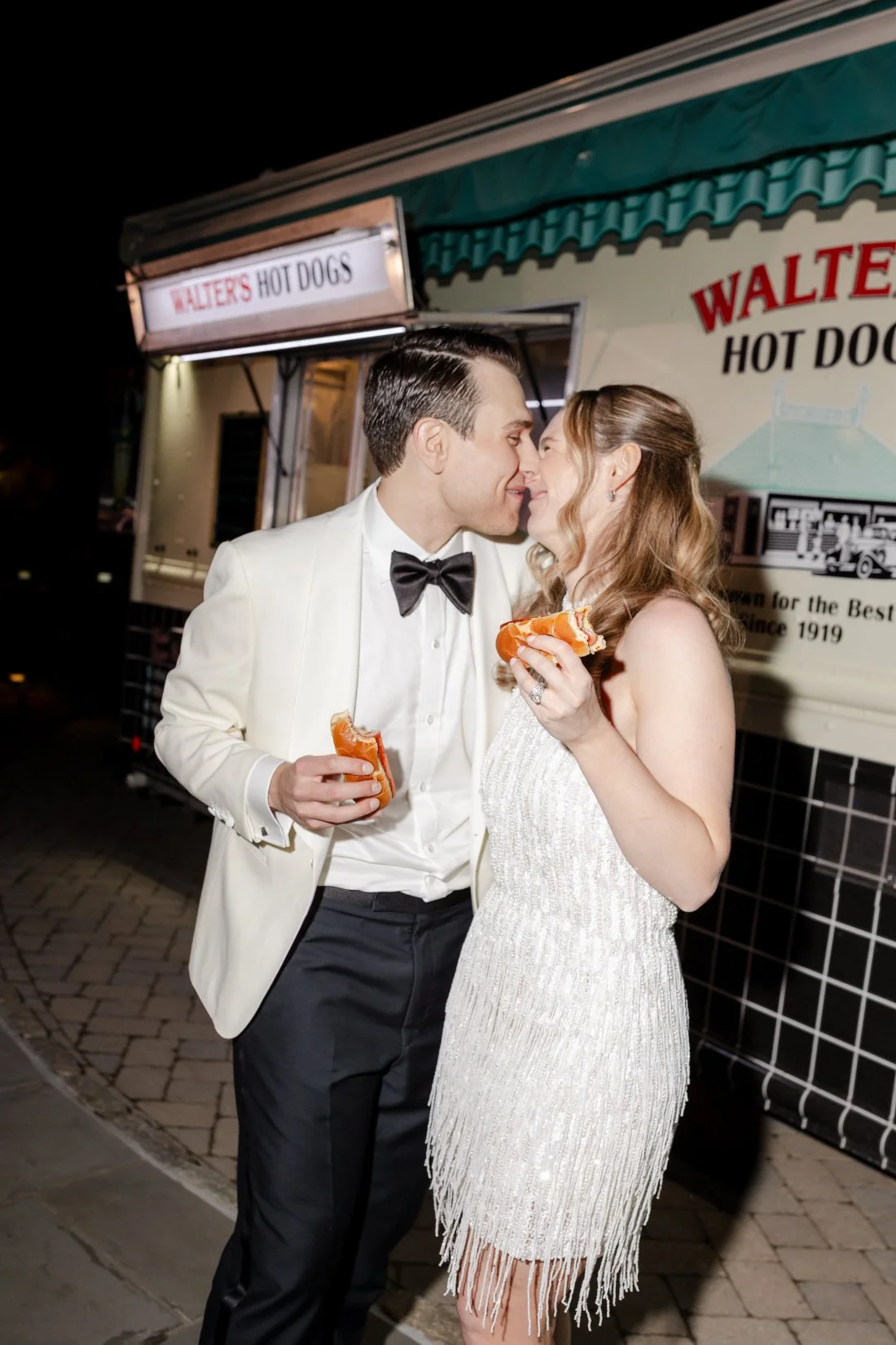 Bride and groom sharing a kiss while enjoying late-night hot dogs.