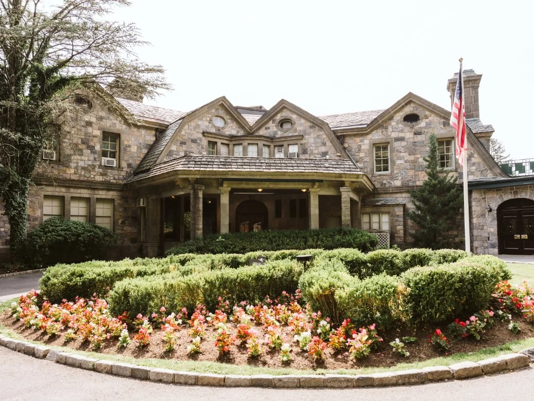Exterior of Tappan Hill Mansion wedding venue in Tarrytown, New York, surrounded by lush greenery and colorful flower beds.