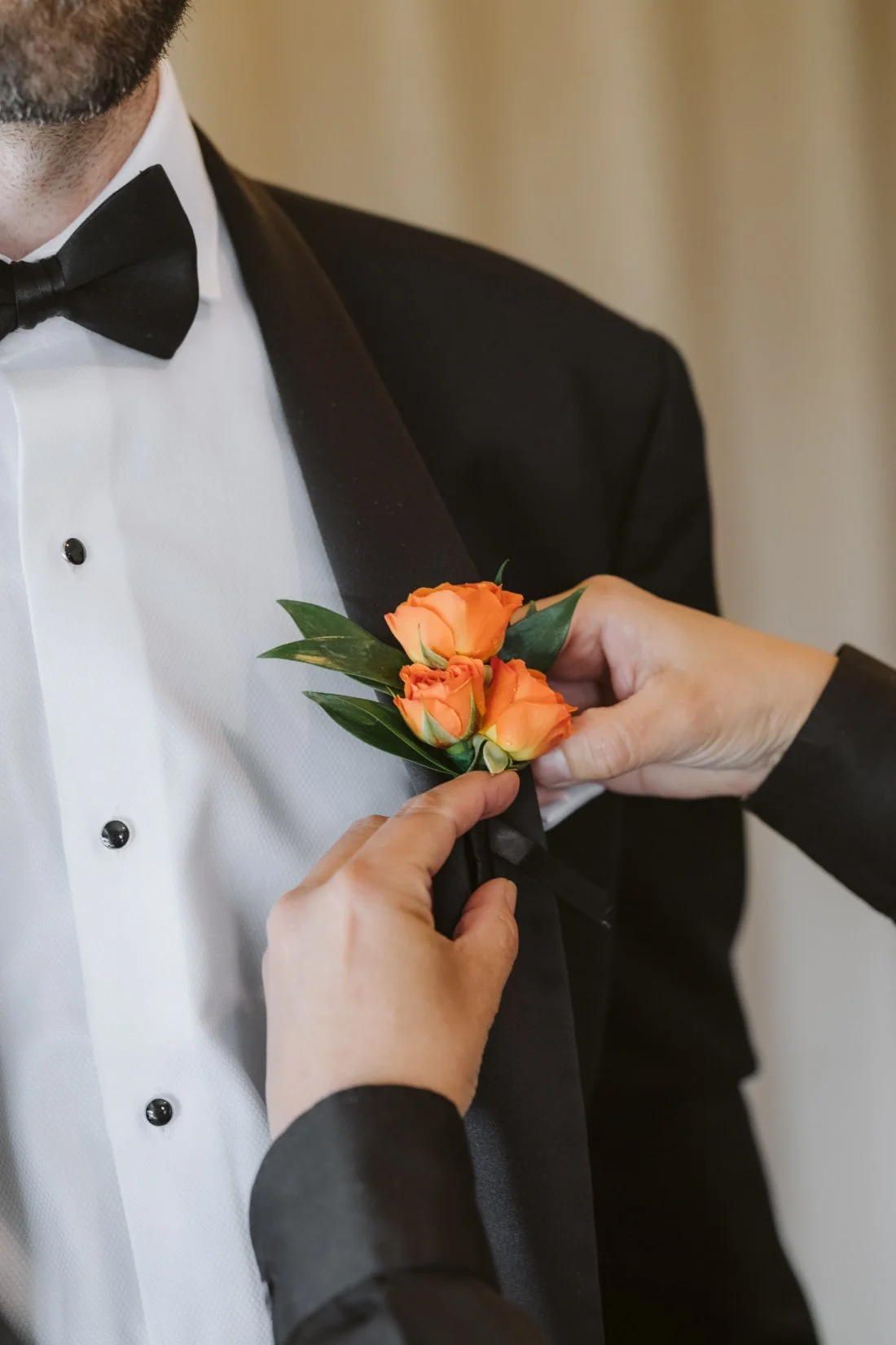 Orange rose boutonniere being pinned onto the groom's black tuxedo jacket before the wedding ceremony.