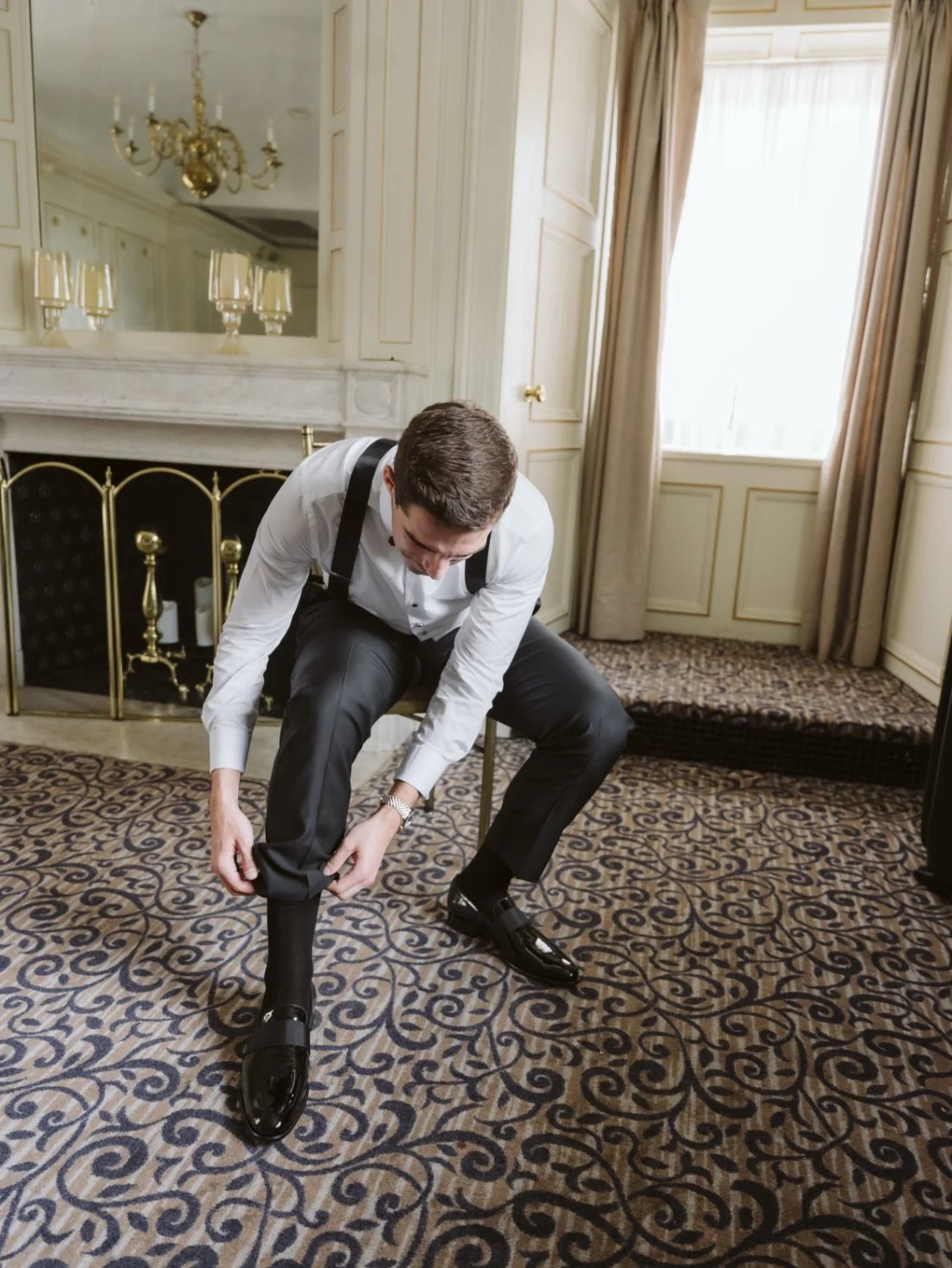 Groom adjusting his black tuxedo pants and polished dress shoes while getting ready for his wedding day.
