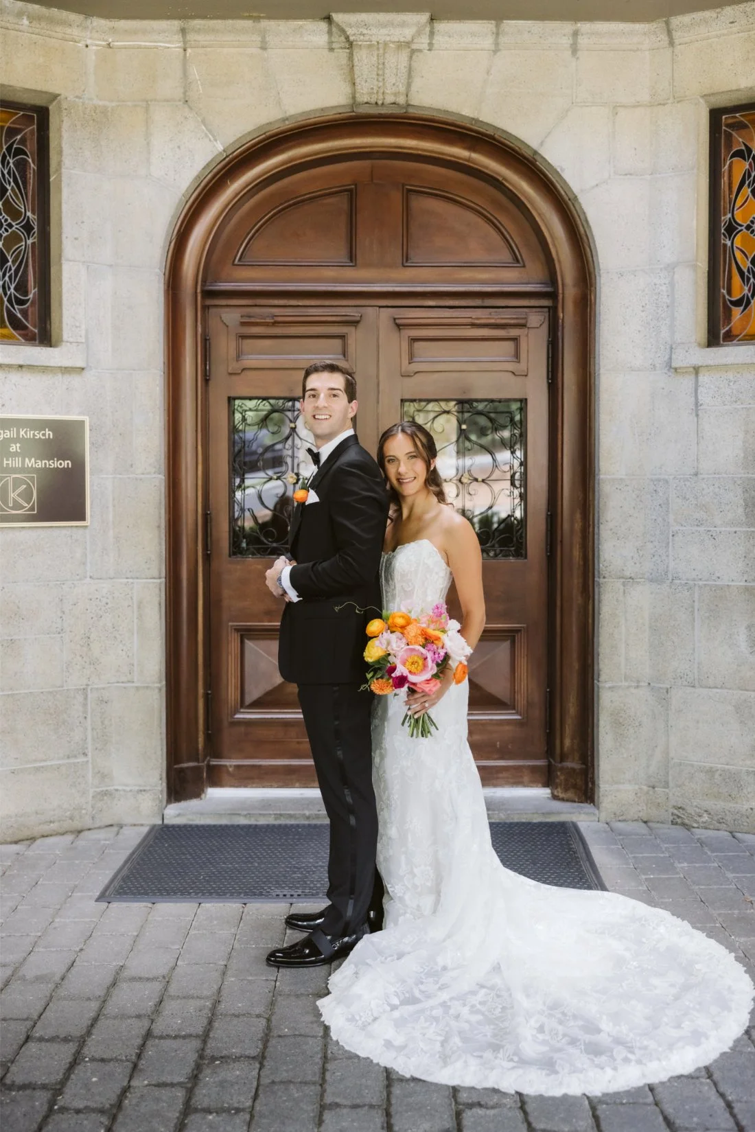 Sydney and Matt pose in front of the grand wooden entrance at Tappan Hill Mansion, showing off her floral lace wedding gown and colorful bouquet.