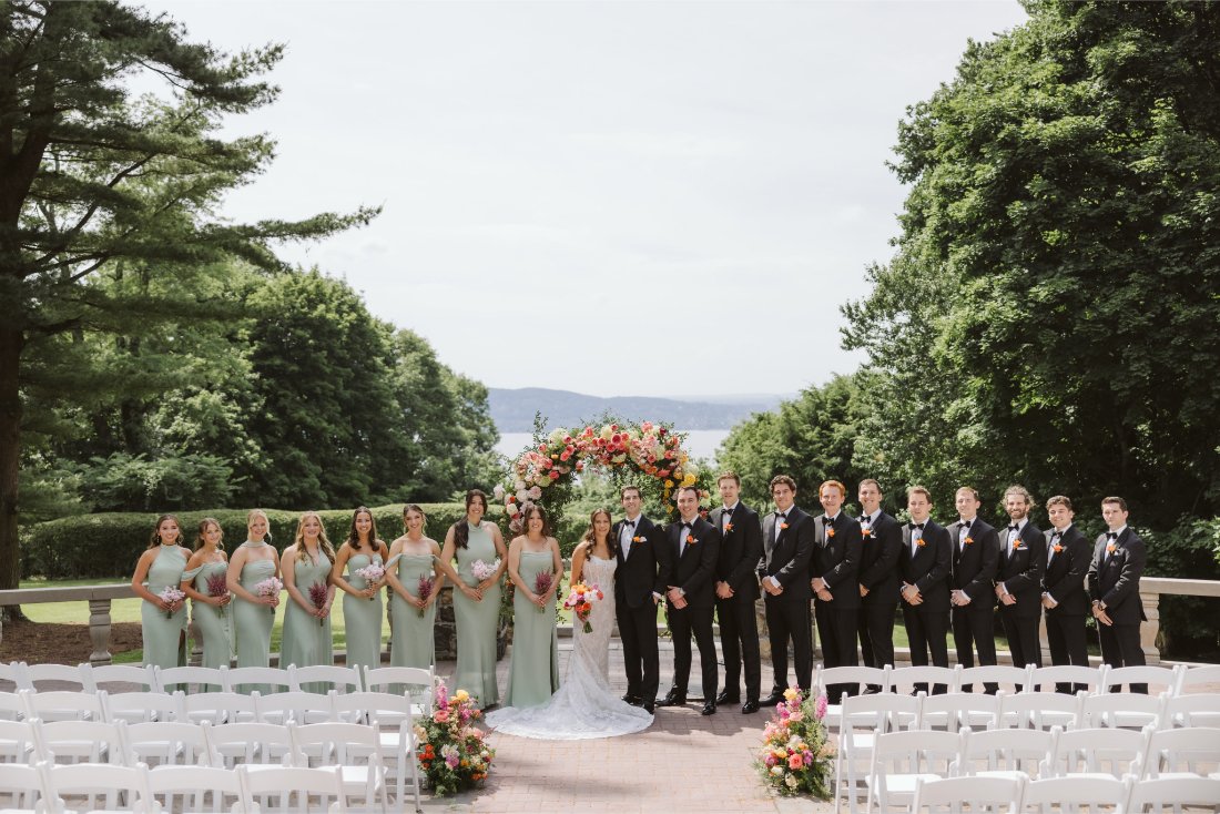 Sydney and Matt pose with their wedding party beneath a colorful floral arch overlooking the Hudson River at their Tappan Hill Mansion wedding.