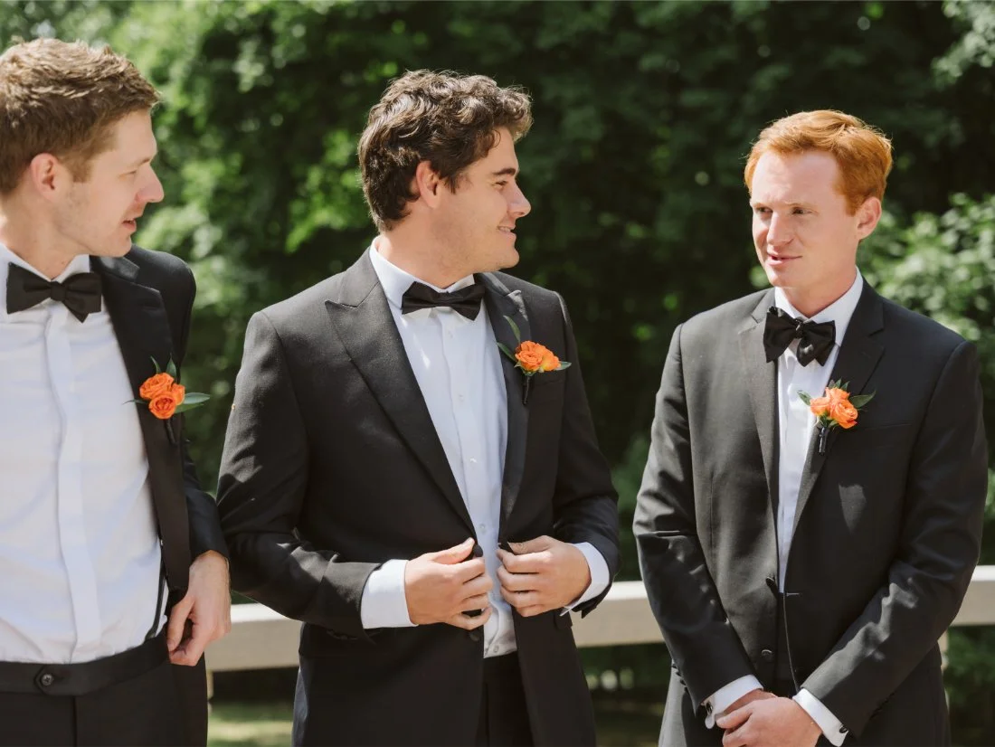 Three groomsmen in classic black tuxedos with bright orange boutonnieres share a moment outdoors before the wedding ceremony.