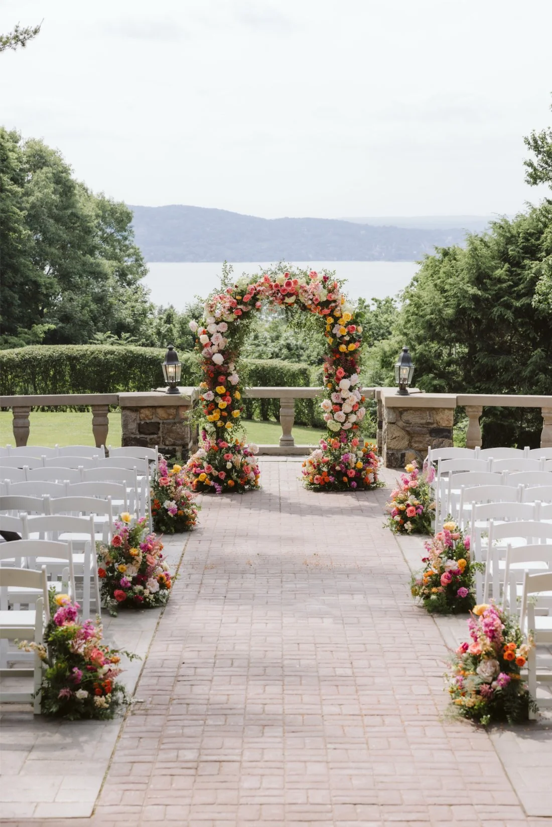 Outdoor Tappan Hill Mansion wedding ceremony setup featuring a colorful floral arch, white chairs, and Hudson River views.