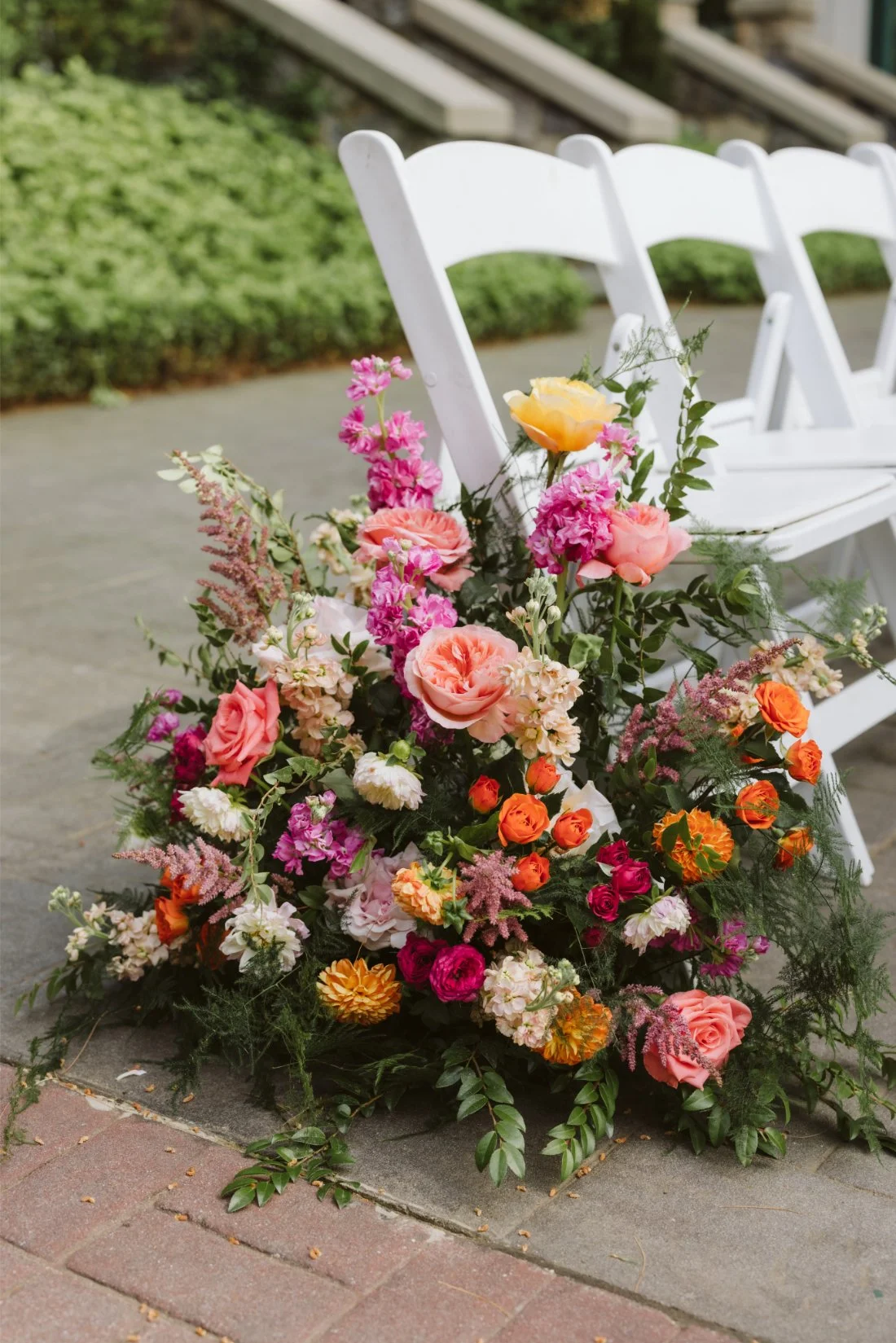 Colorful wedding ceremony flowers featuring pink, coral, orange, and yellow blooms arranged beside white guest chairs.