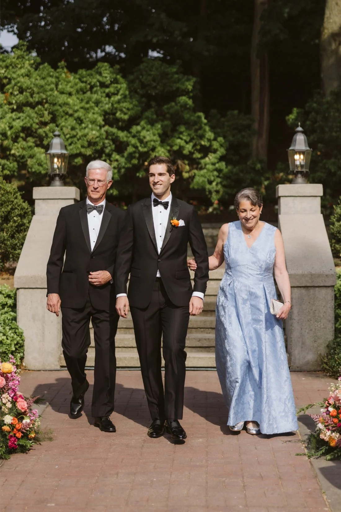 Matt walks down the outdoor wedding ceremony aisle with his parents, wearing a classic black tuxedo with an orange boutonniere.