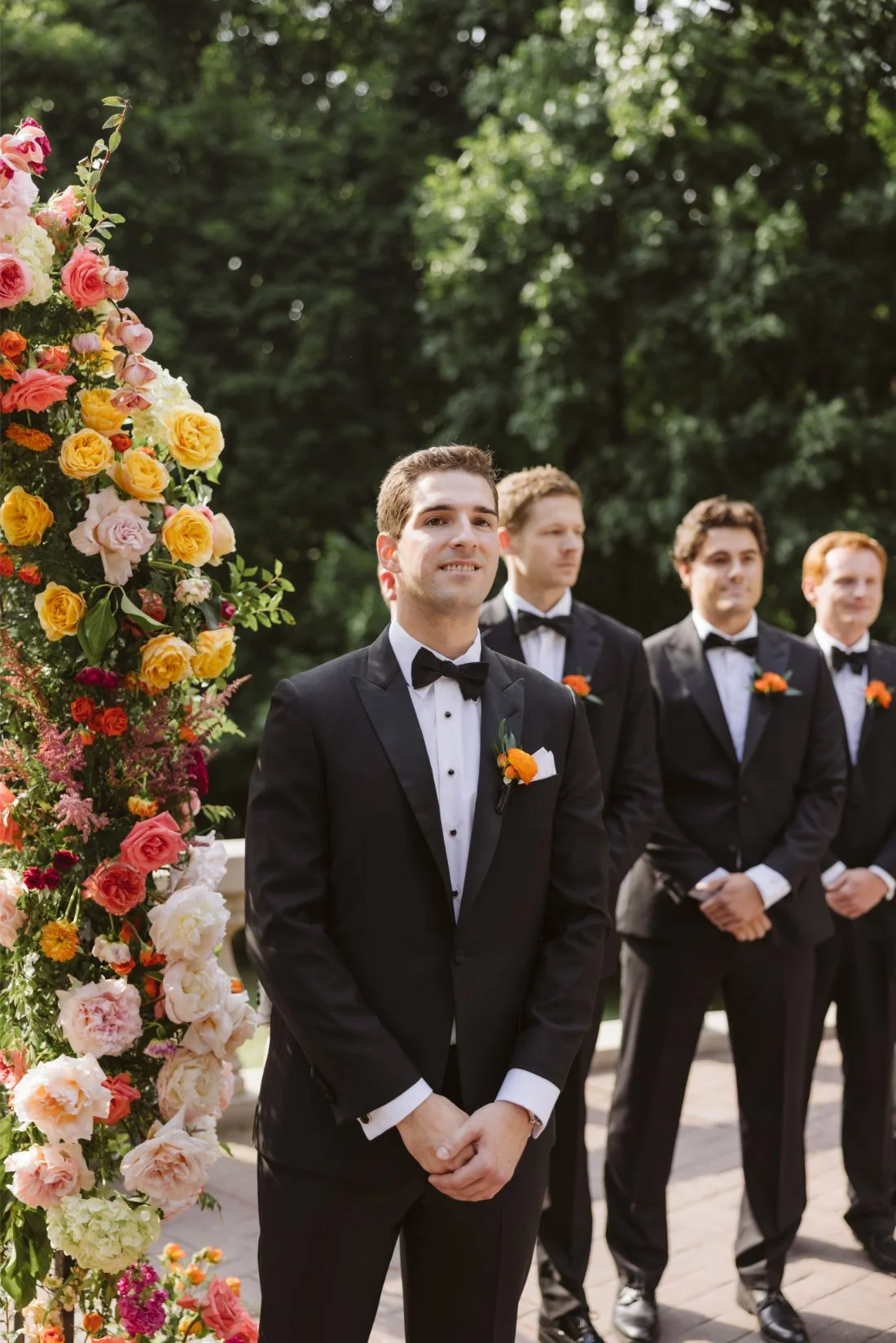 Matt waits at the floral wedding ceremony arch with his groomsmen, wearing black tuxedos with bright orange boutonnieres.