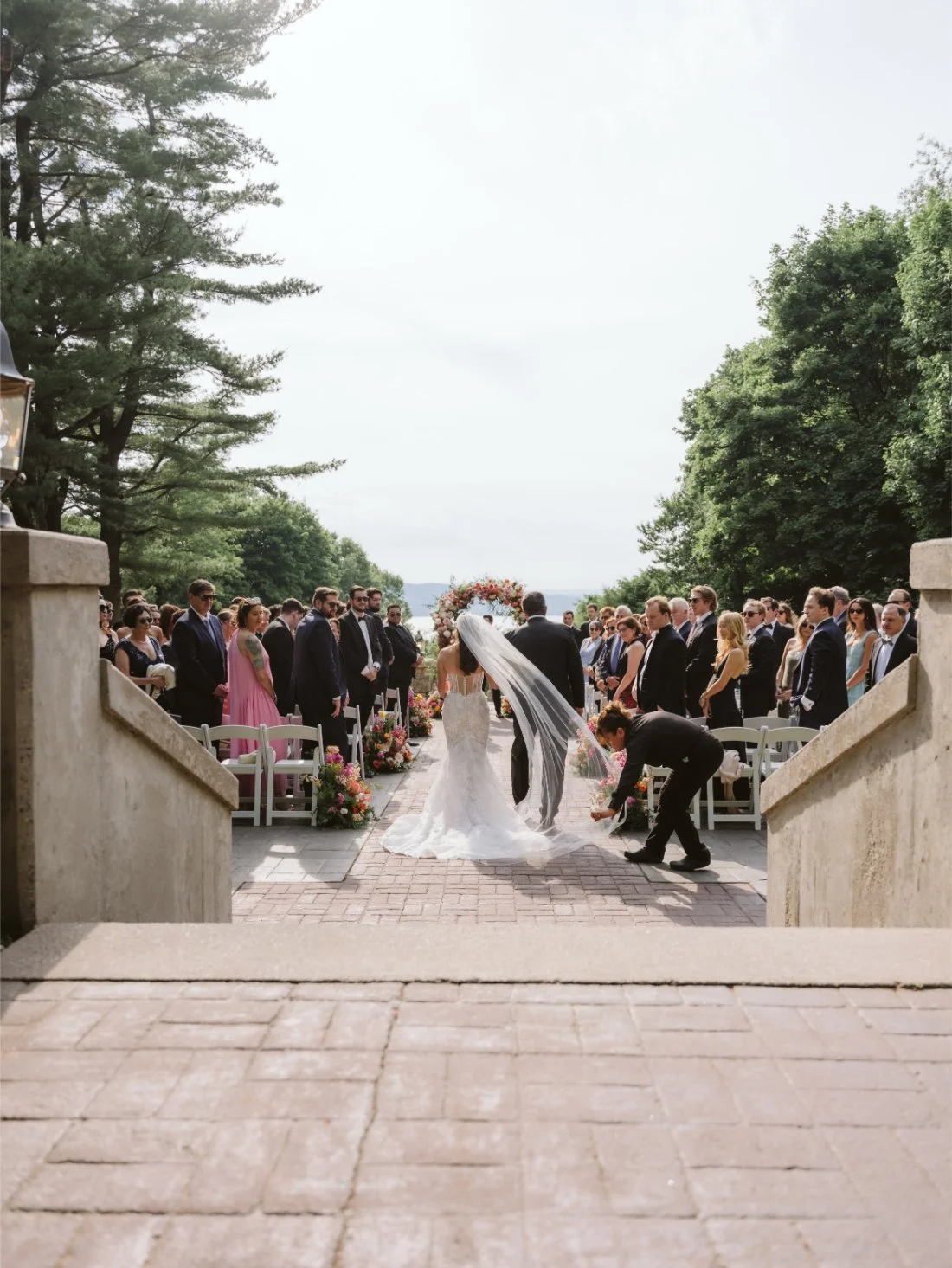 Sydney walks down the aisle with her father at Tappan Hill Mansion as a wedding attendant adjusts her long, flowing veil.