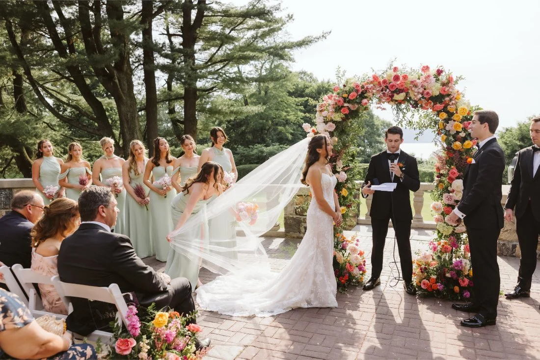 Sydney and Matt stand beneath a vibrant floral arch surrounded by their wedding party and guests during their outdoor ceremony.
