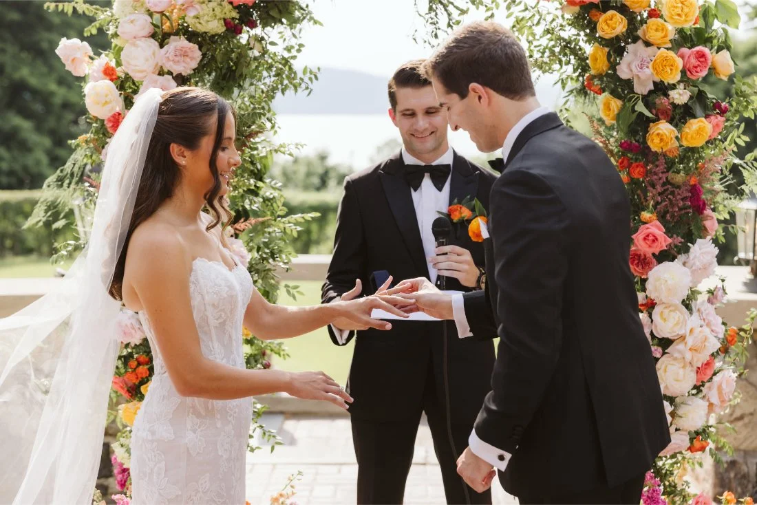 Matt places a wedding ring on Sydney’s finger during their Tappan Hill Mansion wedding ceremony beneath a colorful floral arch.