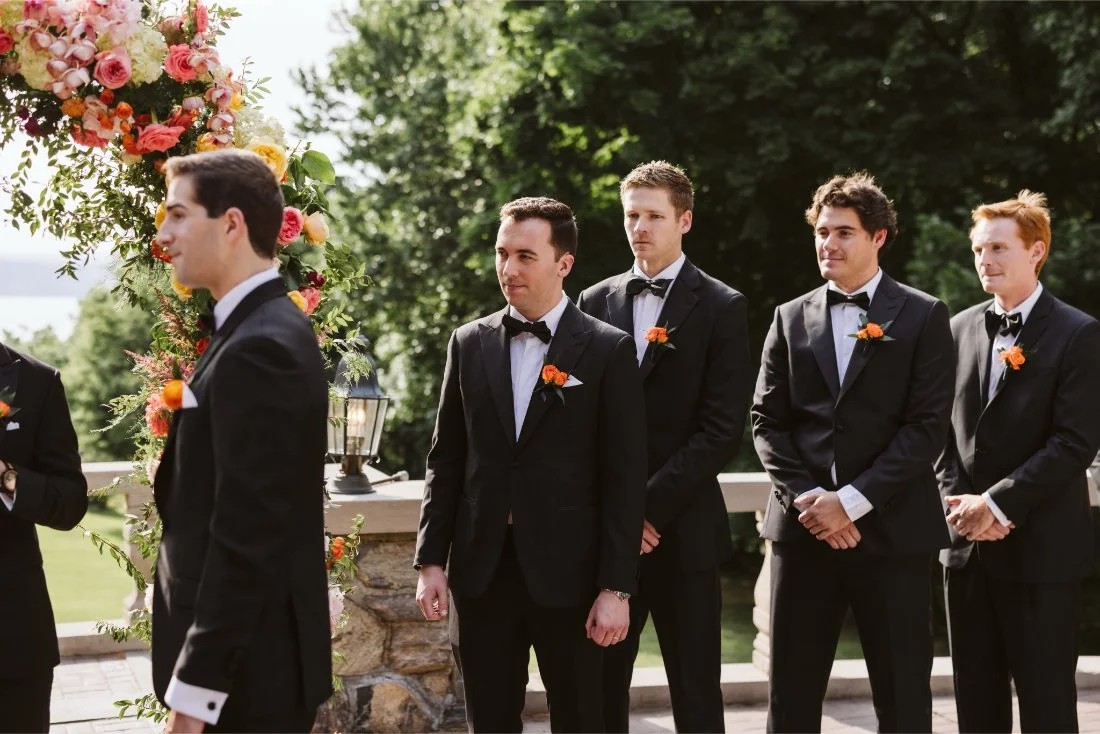 Groomsmen in classic black tuxedos with orange boutonnieres stand beside the floral arch during the Tappan Hill Mansion wedding ceremony.