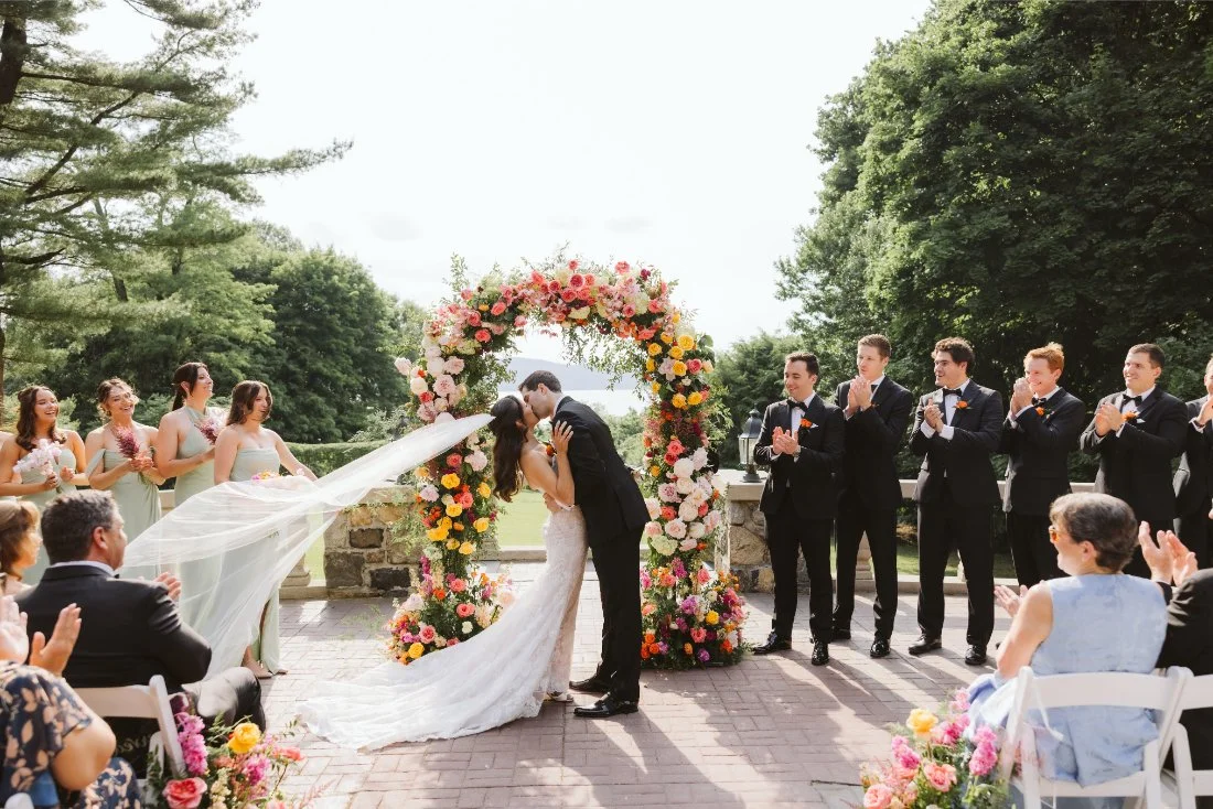 Sydney and Matt share their first kiss as newlyweds beneath a colorful floral arch at their Tappan Hill Mansion wedding.