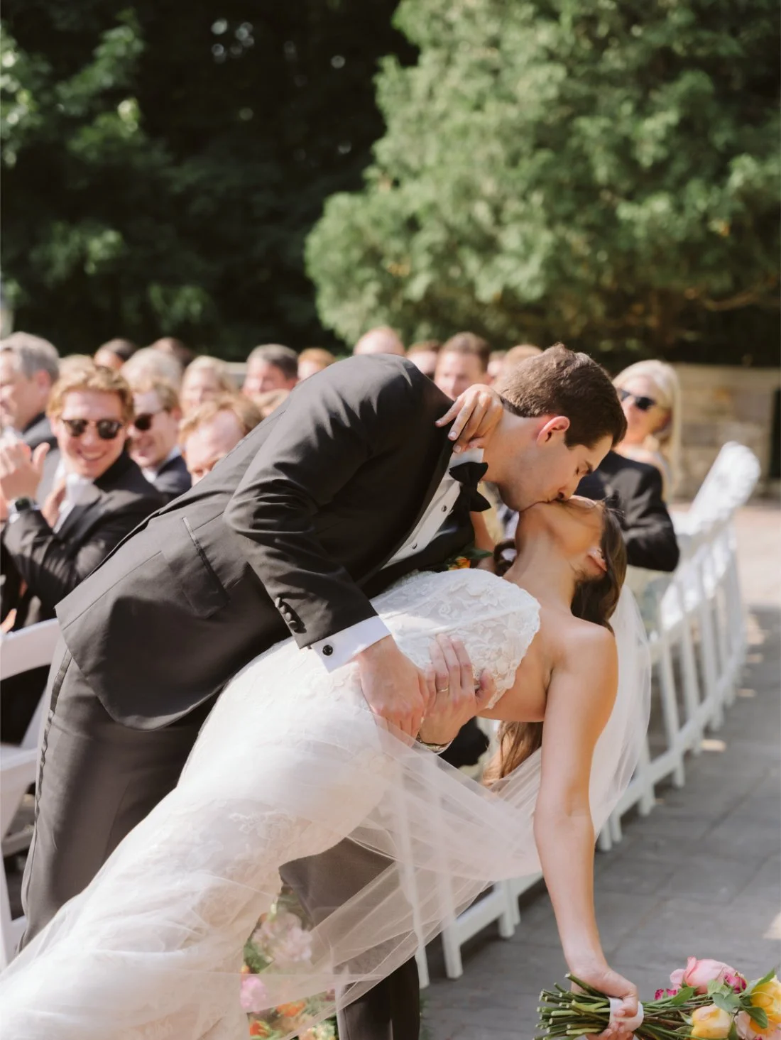 Matt dips Sydney for a romantic kiss after their outdoor wedding ceremony as guests applaud in the background.
