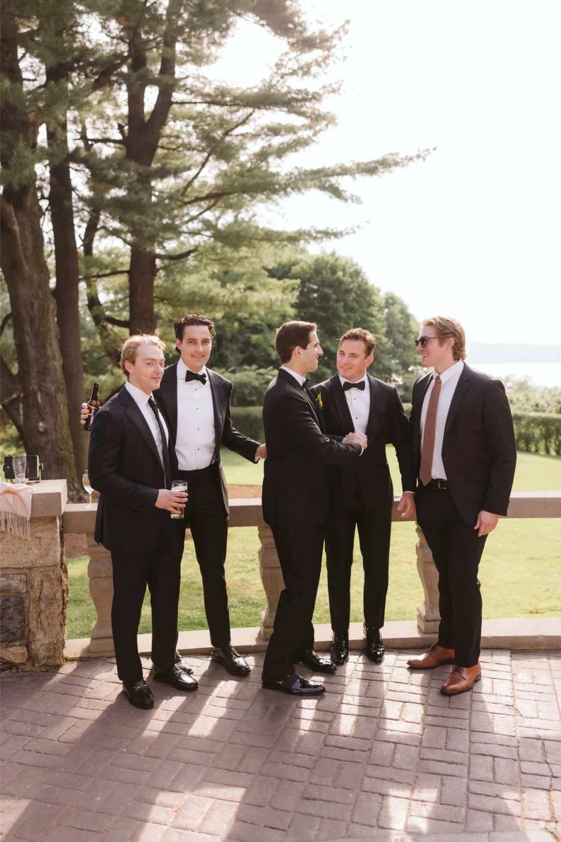 groomsman and groom enjoying cocktail hour on the outdoor terrace at Tappan Hill Mansion, surrounded by lush greenery and scenic Hudson River views.
