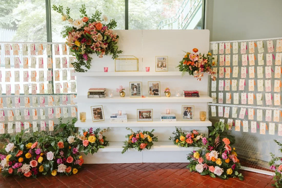 Tappan Hill Mansion wedding escort card display featuring colorful floral arrangements, personalized guest cards, framed photos, and decorative books.