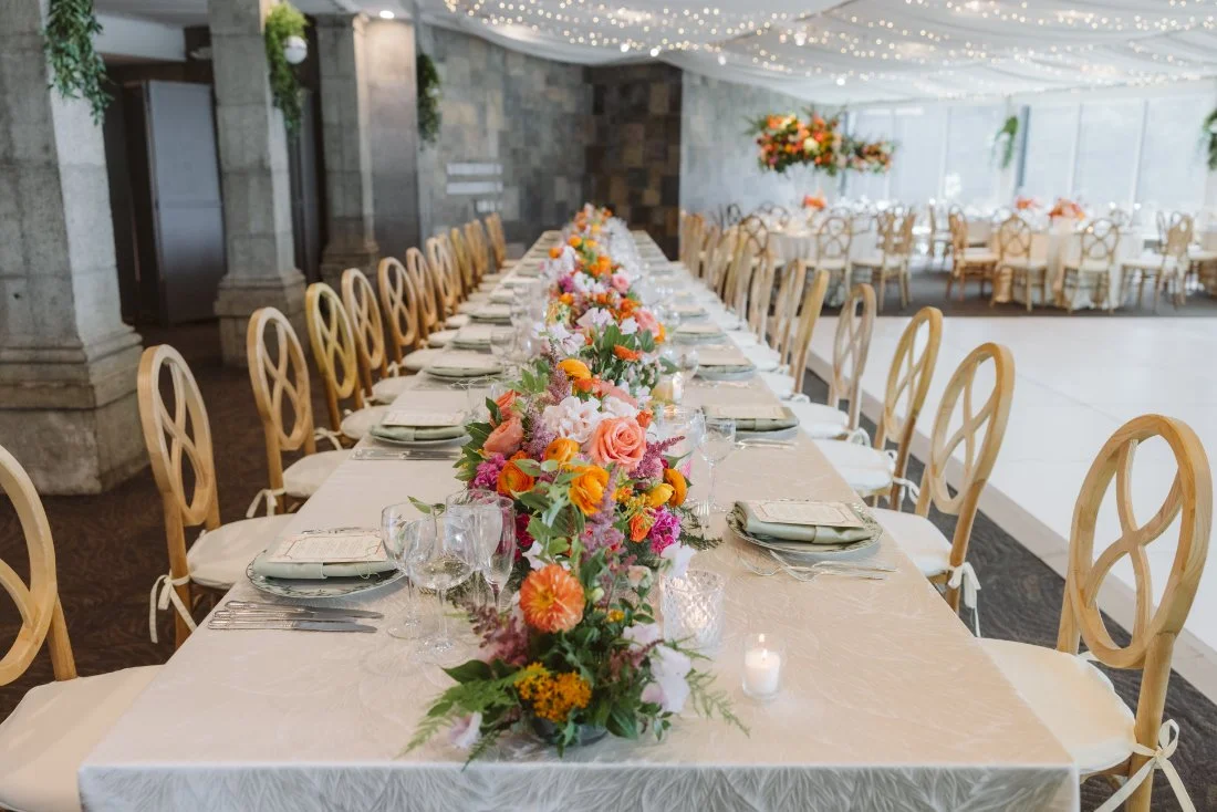 Elegant wedding reception table with colorful garden-style floral centerpieces, sage green napkins, wooden chairs, and twinkling string lights.