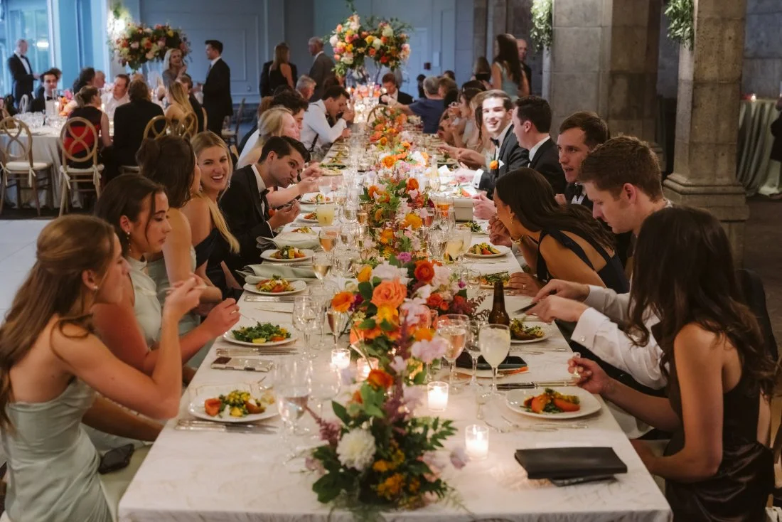 Wedding guests enjoying dinner at a long reception table with colorful floral centerpieces at Tappan Hill Mansion.