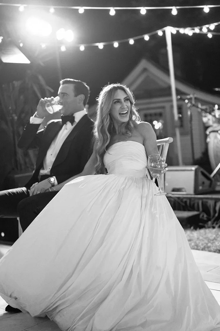 Bride and groom laughing during a reception speech under string lights, capturing a joyful wedding guest experience