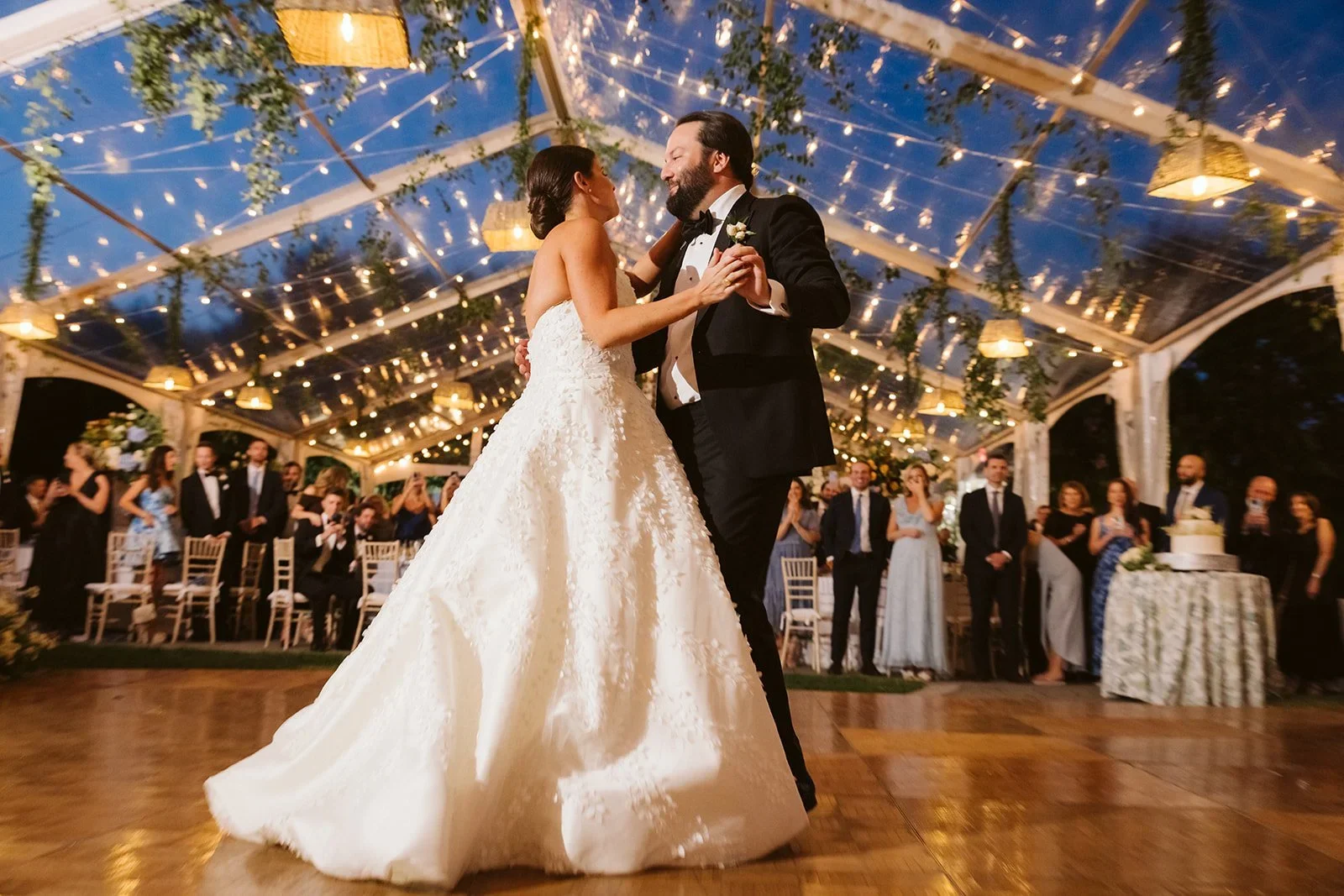 Wedding lighting ideas for a clear-top tent reception featuring warm string lights, hanging greenery, and a couple sharing their first dance.