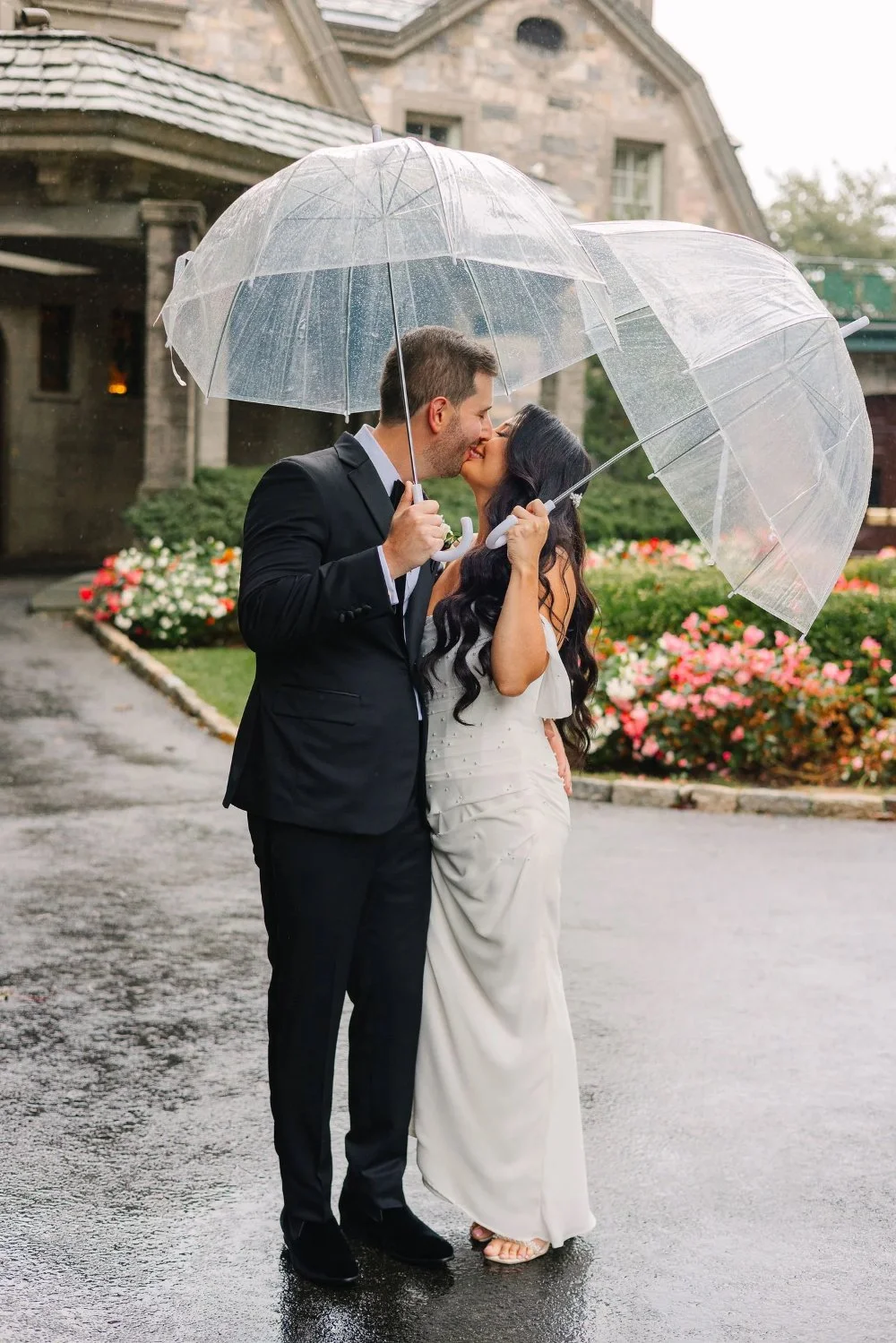 Bride and groom kissing beneath clear umbrellas during romantic rainy wedding portraits outside a stone estate