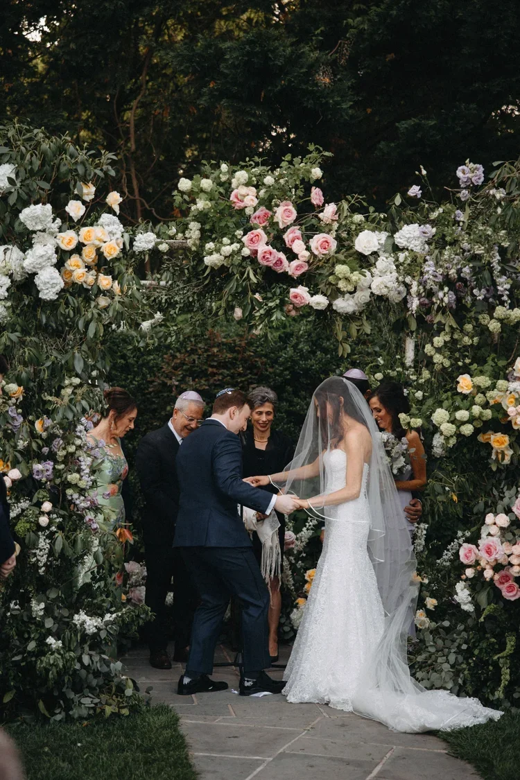 Couple exchanging vows under a lush floral arch outdoors, a perfect example of why weather plans belong on every wedding planner checklist.