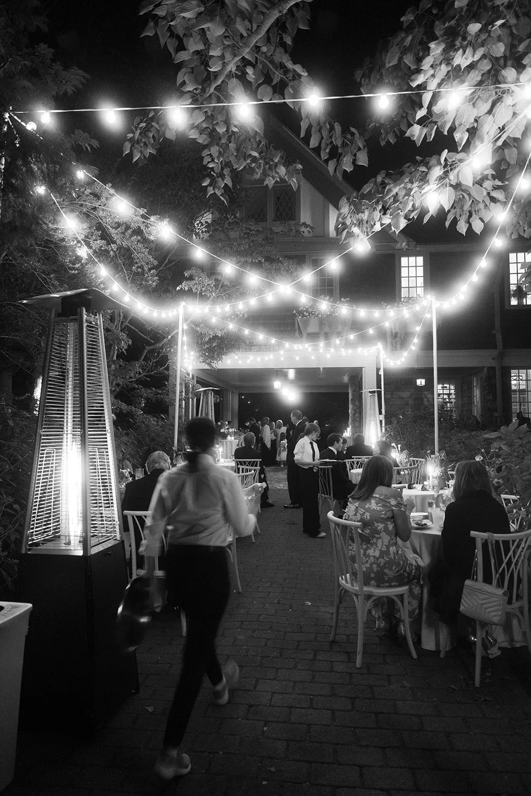 Black and white photograph of an outdoor estate wedding reception with overhead bistro string lights, intimate dining tables, and guests mingling after dark.