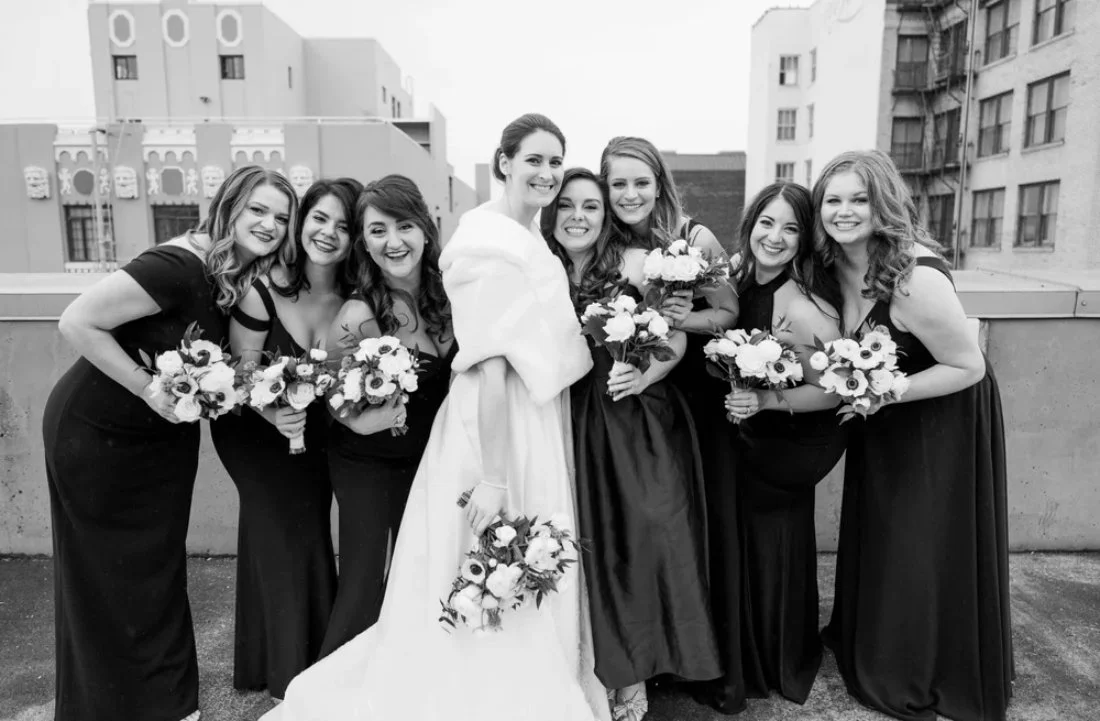 A smiling bride in a white gown and fur wrap stands with her bridesmaids in black dresses on a rooftop, showcasing elegant winter wedding fashion for a bridal party.