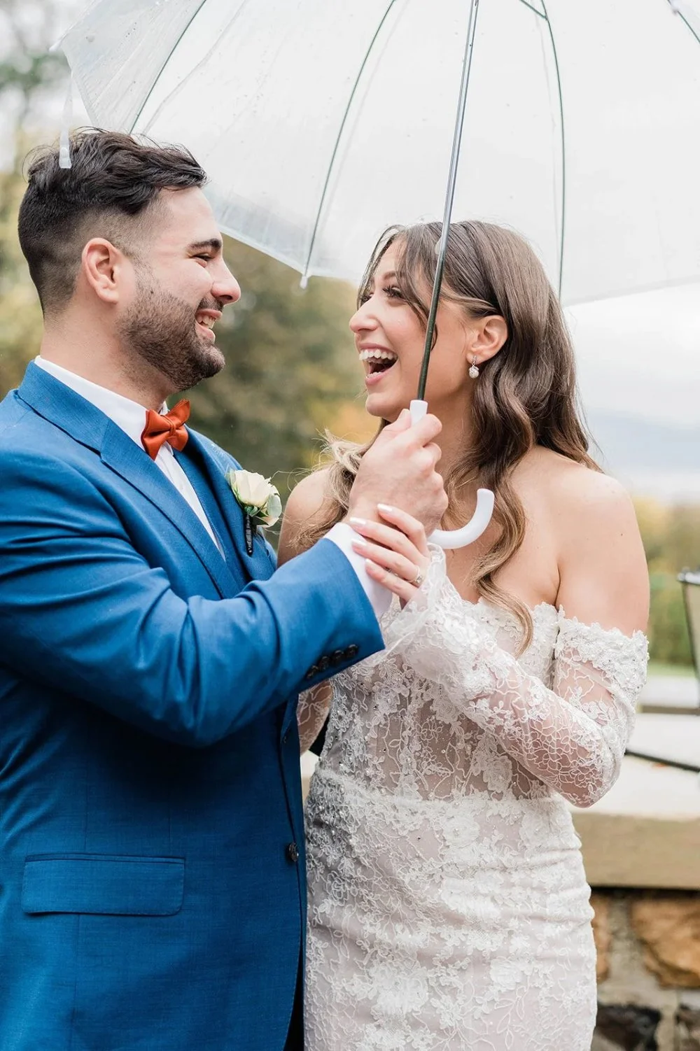 Bride and groom smiling together beneath a clear umbrella during a rainy outdoor wedding portrait